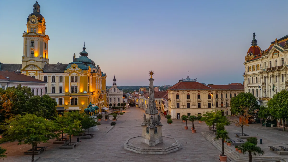 Pécs történelmi Széchenyi tere esti fényekben, középen a Szentháromság-szoborral